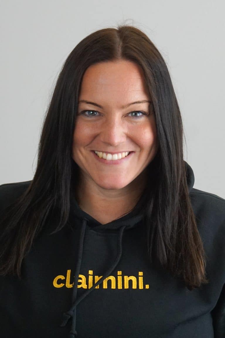 Eine Frau die mittig in die Kamera lächelt mit langen dunklen Haaren und einem schwarzen Hoodie mit gelbem claimini Schriftzug